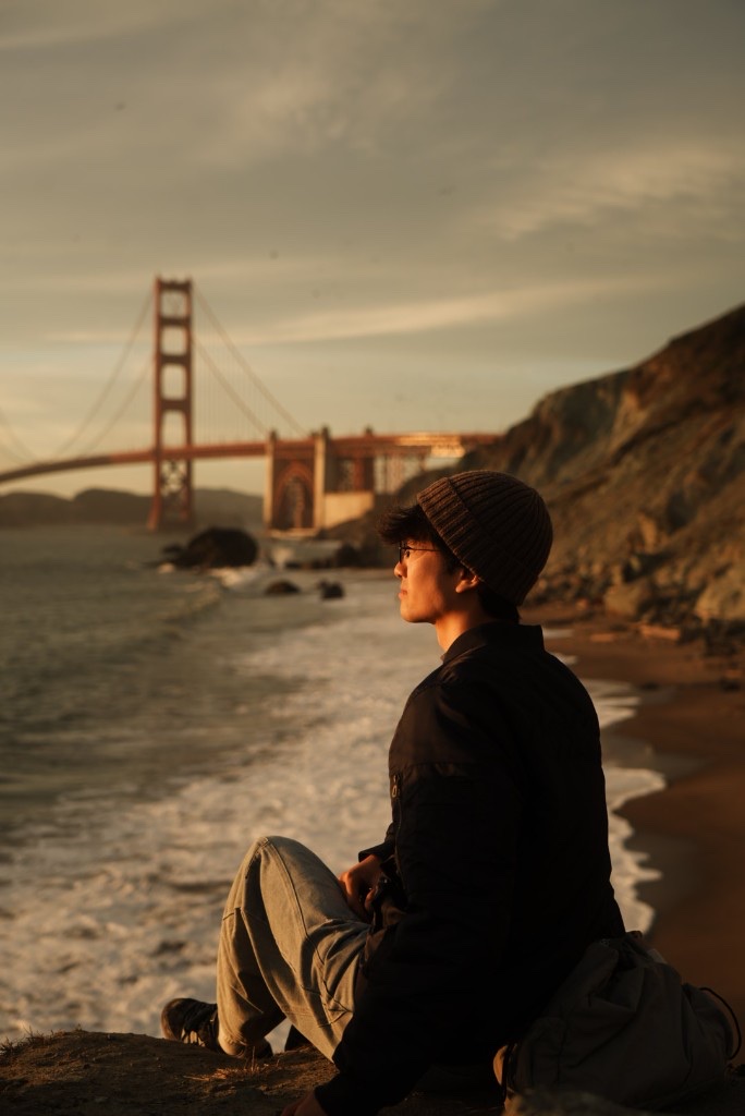 Kayne Duan sitting on a beach, looking toward the Golden Gate Bridge